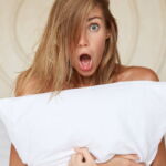 Shocked beautiful young woman with bugged eyes and widely opened mouth, hides nude body with white p