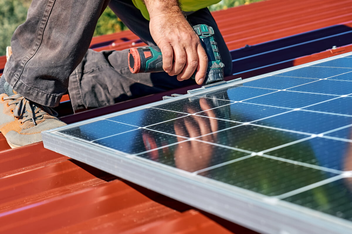 Man's hands with drill installing stand-alone photovoltaic solar panel system on rooftop of a house.