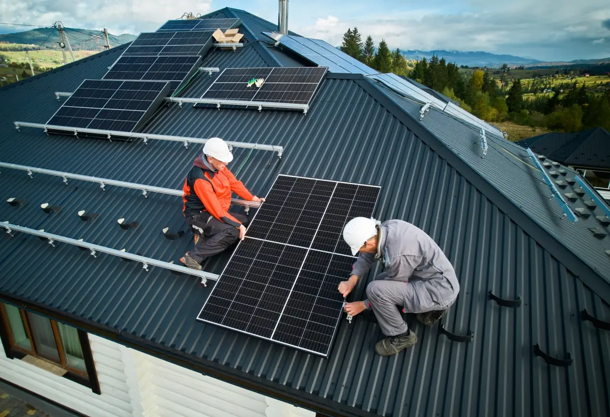 Deux ouvriers en tenue de sécurité installent des panneaux solaires sur le toit métallique d'une maison, présentant un bâtiment photovoltaïque qui soutient la transition écologique au milieu de collines verdoyantes et d'arbres dans un cadre rural.