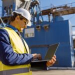 Une personne vêtue d'un gilet de sécurité et d'un casque de chantier utilise un ordinateur portable à l'extérieur, sur un site industriel, où elle surveille les forages au milieu de structures métalliques et d'équipements.