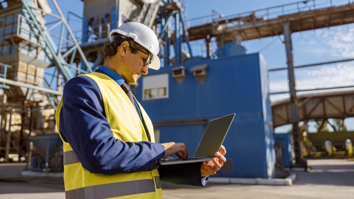 Une personne vêtue d'un gilet de sécurité et d'un casque de chantier utilise un ordinateur portable à l'extérieur, sur un site industriel, où elle surveille les forages au milieu de structures métalliques et d'équipements.